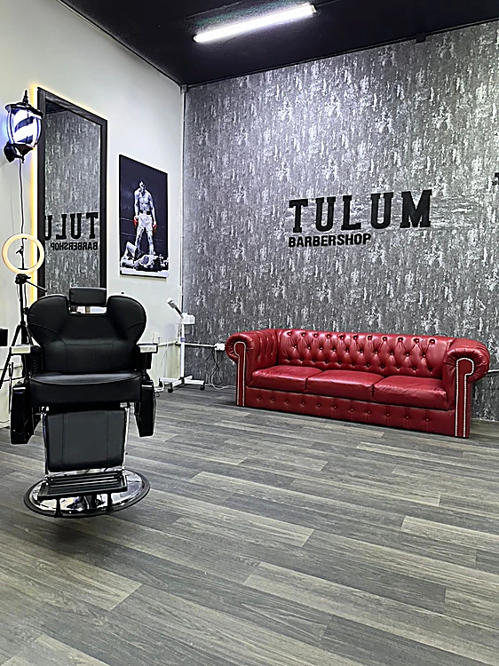 Muro texturizado con el letrero TULUM Barbershop, sillón de barbero y sofá rojo sobre piso de duela gris