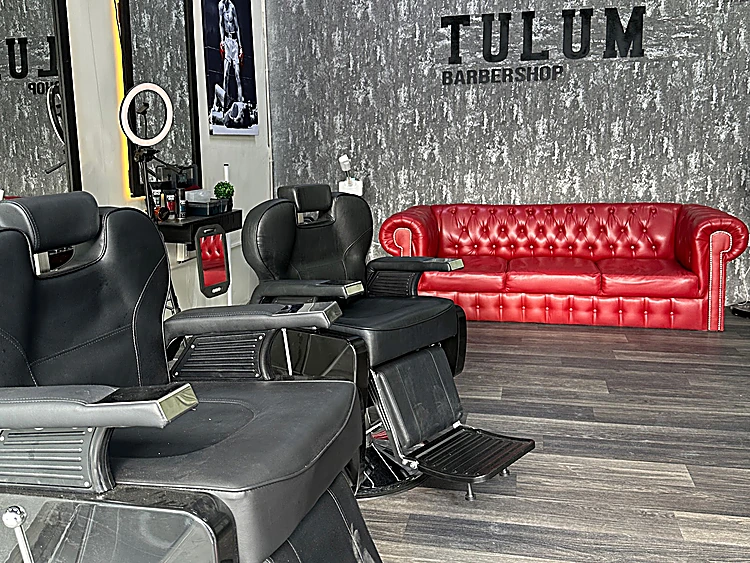 Sala de Tulum Barbershop: letrero TULUM en el muro gris, sofá chesterfield rojo y dos sillones de barbero negros
